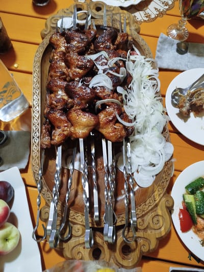 A close-up, top-down shot captures a wooden table set with a meal. In the center, a rustic wooden platter holds numerous skewers of grilled meat, interspersed with thinly sliced white onions. The meat appears to be dark brown and glistening. To the right of the platter, a white plate contains what looks like the remains of a dish, with a few pieces of food and cutlery. Further to the right, a small white plate is visible with chopped cucumbers and tomatoes. In the lower left corner, a white rectangular dish holds two apples and a dark plum. The table surface is made of orange-tinted wood planks. In the background, partially visible, are glasses and other dinnerware. The scene is likely from a restaurant or home dining setting in Kabanbay, Kazakhstan, given the food presentation.