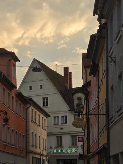 The image shows a tilted view of a street scene in Ansbach, Germany, with buildings of various colors and architectural styles. The sky, filled with soft, cloudy formations, suggests it might be late afternoon or early evening. The buildings are mostly traditional, with pitched roofs and numerous windows. Some buildings have ornate architectural details, while others are more utilitarian.
There are several signs visible on the buildings. One sign, partially obscured, appears to be for "E. KNOBL Gold-schmiede" (Goldsmith). Another red sign reads "DER TOUR". A green neon sign on one of the buildings says "optik.", indicating an optician's shop. The overall impression is that of a historic European town. The scene is devoid of people, focusing solely on the architecture and the sky. The low angle of the photograph, combined with the tilted perspective, creates a dynamic and slightly disorienting view of the urban landscape.