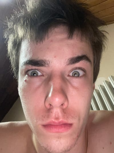 This is a close-up, vertically oriented selfie of a young person. The subject has fair skin with a few small red blemishes visible on their forehead and shoulder. Their dark brown hair is styled messily, appearing somewhat damp and tousled. They have prominent eyebrows and light green eyes that are looking directly at the camera, with a slightly surprised or curious expression. Their lips are parted, revealing pinkness. Light stubble is visible on their chin and jawline. The lighting suggests an indoor setting, possibly during the day, with a bright light source illuminating the subject's face. In the background, a slatted object, possibly a radiator or blinds, is visible. A wooden surface, like a wall or furniture, is also present on the left side of the frame. There is no visible text in the image.