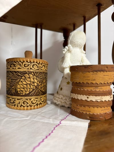The image shows a close-up view of a wooden shelf in a room. On the shelf are two birch bark containers and a crocheted doll. The container on the left is larger and cylindrical, with intricate carvings of pinecones and swirling patterns. It has a wooden knob on top. To the right of it, there are two smaller stacked birch bark containers with decorative scalloped edges and ribbon accents. Behind these objects, a white crocheted doll with a conical head and ruffled clothing is partially visible. The shelf itself appears to be made of dark wood, and the background is mostly a plain white wall with some indistinct wooden elements on the right. The items are placed on a white cloth with a visible pink embroidered edge. The image does not provide any information about the city of Ust'-Kabyrza, Russia, or any specific context related to it.