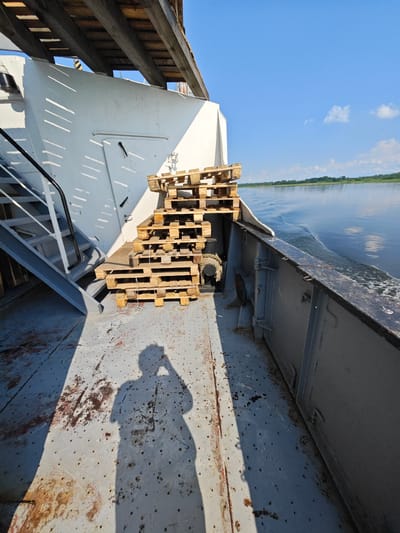 The image shows a side view of a ferry navigating a wide, calm river under a bright blue sky with scattered clouds. The ferry deck is made of weathered grey metal with some rust stains and scattered dark spots. To the left, a grey metal staircase with white railings leads upwards. A stack of wooden pallets is positioned on the deck, to the right of the stairs. The ferry's white superstructure is visible, with slatted shadows cast across it. In the foreground, a shadow of a person, holding what appears to be a camera or phone, is cast on the deck. The river is tranquil, with gentle ripples and a wake trailing the ferry. The far bank of the river is lined with trees, indicating a rural or semi-rural setting. The location is identified as Novonazimovo, Russia.