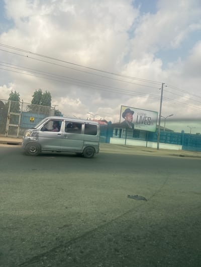 The image presents a street scene in Elimgbu, Nigeria, under a partly cloudy sky. A silver-grey minivan, exhibiting an unpainted front left fender area and a darker, potentially smaller rear wheel, traverses the asphalt road from left to right. Three occupants are visible within the minivan: a driver wearing a red and white striped shirt, and two passengers in the rear.

On the right side of the road, a large billboard displays a portrait of a man in a dark hat and suit, with the prominent text "A LEGEND LIVES ON" and the dates "1959 - 2019". Behind the billboard and further left, structures are delineated by blue painted fences and walls. To the far left, a grey metal fence topped with barbed wire encloses an area featuring green trees. Multiple overhead utility lines cross the sky, supported by a dark utility pole positioned near the billboard. The asphalt road surface in the foreground is visible, with a dark, flat piece of debris present in the lower right quadrant.