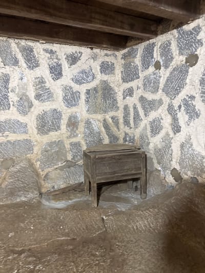 The scene depicts the interior of a structure, likely a building or cave, in the Sà Phìn Commune of Vietnam. The walls are constructed of rough-hewn stones, arranged in an irregular pattern. A small, wooden box is affixed to one of the walls. A wooden beam is partially visible. The environment appears dark and possibly damp.