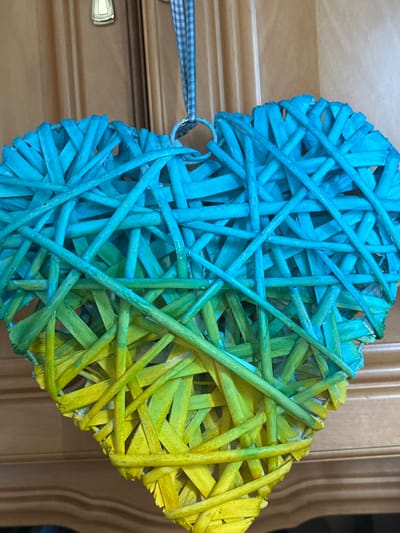 A close-up, slightly angled shot captures a large, heart-shaped decoration made of woven reeds or twigs. The decoration is suspended by a blue and white checkered ribbon attached to a loop at the top. The heart exhibits a gradient of colors, transitioning from a bright, vibrant blue at the top to a rich green in the middle, and finally to a sunny yellow at the bottom. The woven texture of the material is clearly visible, with many individual strands crossing and interlacing. The background consists of a wooden surface, likely a door or cabinet, with visible grain and a horizontal line suggesting paneling. A small, brass-colored handle or latch is visible on the wooden surface above and slightly to the left of the heart decoration. The image does not depict any people or specific urban landmarks in Bad Blankenburg, Germany.