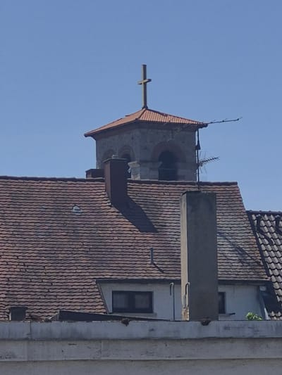 A close-up, eye-level shot captures the upper portion of a building, likely a church or a historical structure, against a clear blue sky. The building features a red-tiled roof with visible shingles. A prominent feature is a bell tower with a copper-colored roof, topped by a golden cross. The tower has arched openings and stone detailing. Attached to the tower is an antenna, suggesting the building is also used for communication purposes. In the foreground, a portion of another building with a white facade and windows is visible, also with a red-tiled roof. A tall, rectangular concrete structure, possibly a chimney, stands in front of the bell tower. The overall scene suggests an urban or semi-urban environment in Bruchsal, Germany.