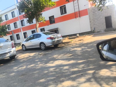 A street view in Abay, Kazakhstan shows a silver Peugeot 408 sedan parked on a dirt and gravel surface. The car, license plate "580 ADN 09," is facing left. To the left of the Peugeot, a white Chevrolet sedan is partially visible, facing left, with its license plate showing "63 AAG 09." Behind the cars, a multi-story building features alternating horizontal bands of white and orange siding. A metal fire escape ladder is attached to the building's facade. To the right of the building, a section of light-colored brick wall with a black door and concrete steps is visible. Trees with green foliage are present, casting shadows on the ground. The sky is clear and blue. The image is captured from a low-angle perspective, possibly from inside another vehicle, as a side mirror is visible in the lower right corner.