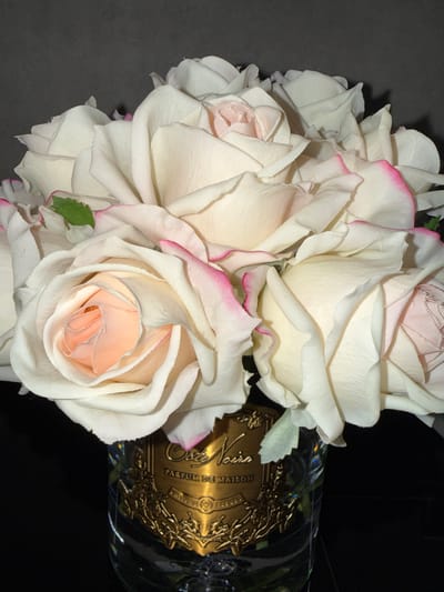 An eye-level shot captures a vibrant bouquet of artificial roses alongside a decorative bottle, set against a dark, featureless background, possibly a dark tabletop or wall. The image is oriented such that the roses are on the left and center, and the bottle is on the right, appearing to lie horizontally.

The subjects of the image are a cluster of fabric roses and a clear glass bottle with an ornate golden label. The roses are primarily white and cream, with many petals featuring distinct pink edges, while the inner petals of some blooms are a soft peach or light pink. Their fabric texture is visible, indicating they are artificial. The arrangement is dense, showcasing multiple blossoms in various stages of opening.

Adjacent to the roses, a clear glass bottle is partially visible. It features a highly detailed, embossed golden label. The label reads "MARIE DU VALOIS" across the top, with "PARFUM DE MAISON" directly below it. In a prominent, larger script, the word "Noire" is visible, and the word "FLEURS" also appears on the label. The intricate design includes what appears to be a crest or stylized emblem with decorative flourishes.

The scene suggests a still-life arrangement, possibly for display or photography. There is no discernible activity taking place. The lighting is focused, highlighting the colors and textures of the flowers and the metallic sheen of the bottle's label, creating a dramatic contrast with the dark background. No specific time of day or weather is indicated. The image does not provide explicit visual cues to confirm the geographical location of Zvezditsa, Bulgaria.
