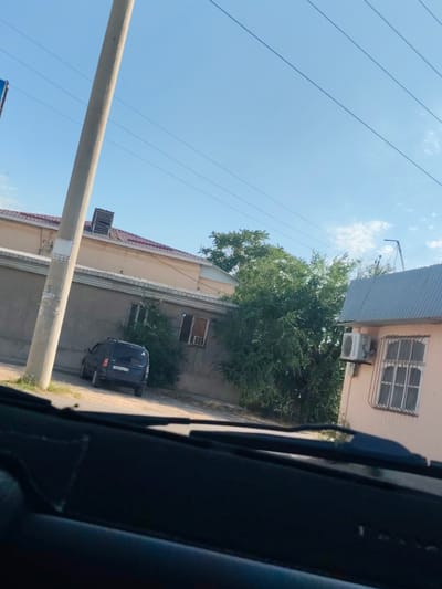 The image depicts a scene viewed from inside a vehicle, likely parked on a street in Şardara, Kazakhstan. The perspective is from the driver's seat, with the dashboard partially visible in the foreground.
The main subjects include a dark-colored minivan parked on a dirt road to the left of the frame. Behind the car, a long, light-colored building with a reddish-brown tiled roof and several windows is visible. A tall, gray utility pole stands prominently in the foreground, positioned between the vehicle and the building. Further to the right, another building with a light-colored facade and a corrugated metal roof is present. This building has a window with a metal grate and an air conditioning unit mounted on its wall. Dense green trees are situated between the two buildings, obscuring parts of them and adding a natural element to the urban landscape. Power lines stretch across the bright blue sky, indicating an outdoor setting.
The activity appears to be a pause or stop, with the car parked and no people visible. The time of day is clearly daytime, suggested by the bright sky and clear visibility. The weather seems fair and sunny. The presence of the utility pole, buildings, and parked vehicle suggests a residential or commercial area.
Visible text includes a license plate on the minivan, which reads "241 AIC 13". There is also some text on a sticker attached to the utility pole, but it is too small and blurry to be deciphered. The overall impression is a quiet, ordinary day in a somewhat dry, sunny environment.