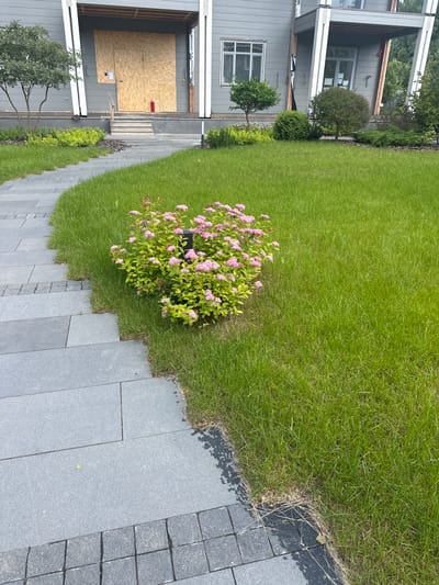 The image depicts a paved pathway leading to the entrance of a modern house. The pathway is constructed with large, rectangular gray stone slabs, bordered by smaller, darker gray square stones. To the right of the pathway is a lush green lawn. In the foreground, a bush with small pink flowers is visible. The house, painted in gray, features large windows and a porch area covered with plywood, suggesting ongoing construction or renovation. A red fire extinguisher is placed near the entrance. The environment appears to be an outdoor residential setting, likely in a suburban area. The city of Uyut, Russia, is not visually identifiable within the image.