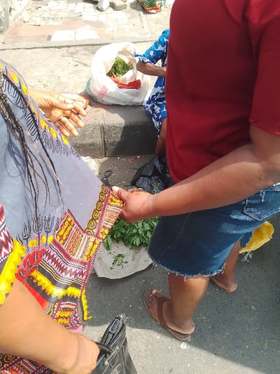 This outdoor scene, set in Mgbuoshimini, Nigeria, captures an interaction at what appears to be a roadside market stall on a bright, sunny day.

Two people are prominently featured in the foreground. On the left, a person is partially visible, wearing a gray top adorned with vibrant yellow, red, and black African print patterns on the lower part and sleeves. Dark braids fall over their left shoulder. Their right hand is extended, palm up, revealing a ring on one finger, while their left hand holds a black handbag, partially visible at the bottom left.

Facing them, a person on the right, also partially cropped, wears a red t-shirt and blue denim shorts with a frayed hem. They have brown thong sandals on their feet. Their left hand is holding onto the sleeve of the person on the left's garment, suggesting a close interaction, possibly a negotiation or transaction.

In the background, behind the interacting individuals, market produce is displayed. A white sack contains green leafy vegetables and some red items, possibly chilies or peppers. Another pile of fresh green leafy vegetables rests directly on the ground or a mat in front of the right person. Black plastic bags are visible on the ground near the dark gray concrete curb, and a yellow plastic bag is near the right person's feet. A third individual, wearing a blue and white patterned garment, is partially seen in the background, seated or kneeling beside the produce. The ground consists of concrete paving, indicative of an urban or semi-urban outdoor setting. No discernible text is visible in the image.