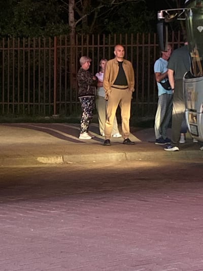 In Terespol, Poland, at night, a group of people are gathered on a sidewalk next to a fence and what appears to be a bus. A man in the foreground, wearing a tan jacket and pants with a black shirt, stands facing the camera. To his left, a woman in a patterned jacket and pants, and another woman in a white shirt and light-colored pants are visible. To the right of the man in tan, two other individuals are partially obscured by the bus. The lighting suggests it is dark, with artificial light illuminating the scene from the front. The ground is paved, and the fence is made of vertical metal bars.