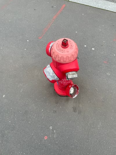 The image displays an overhead view of a red fire hydrant positioned on a dark gray asphalt surface. The hydrant features a ridged, pinkish-red top cap inscribed with the text "YACO," "100," and "5 3/4 TG 1978," and is equipped with a square operating nut. The main body of the hydrant is red, exhibiting areas of worn silver-colored paint or material around its upper and mid-sections. White graffiti is visible on the lower red portion of the hydrant. Two outlets are present; one is a larger, capped port, while the other is a smaller, uncapped port with a white interior, showing wear and exposure. The surrounding asphalt pavement contains a distinct diagonal red line marking in the upper left quadrant and a faint small red circular marking near the lower left. White specks are distributed across the asphalt, which also shows dark, irregular moisture or stain marks immediately around the base of the hydrant. A lighter gray concrete curb or sidewalk edge is visible along the upper right perimeter of the scene. This location is in Montigny-le-Bretonneux, France.