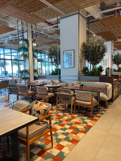 This media file depicts the interior of a modern, stylish cafe in Ras Al-Khaimah, United Arab Emirates. The scene is set during daylight, with natural light streaming in through large windows, suggesting a bright and airy atmosphere. The cafe's design is contemporary and chic, featuring a mix of materials and textures.
The primary subjects are the furnishings and the overall ambiance of the space. We see an array of comfortable seating options, including sofas and chairs with light-colored upholstery and wooden frames. Several chairs are adorned with colorful throw pillows featuring geometric patterns. Tables are also visible, some set with small decorative items.
The setting is characterized by a vibrant, geometric tiled floor in shades of red, beige, and teal, creating a striking visual contrast with the neutral tones of the furniture. In the background, large windows offer glimpses of greenery and trees, suggesting the cafe might be situated near an outdoor area or a landscaped space. Hanging plants and decorative trees are strategically placed throughout the interior, adding a touch of nature and freshness to the environment.
The cafe appears to be unoccupied by people, giving it a serene and polished feel. The overall mood is tranquil and inviting, ideal for relaxation or dining. No visible text is present in the image, contributing to a clean and uncluttered aesthetic. The high ceilings and open layout further enhance the sense of spaciousness. The use of natural materials like wood and woven textures, combined with the modern furniture and colorful tiles, creates a unique and appealing aesthetic for the cafe.