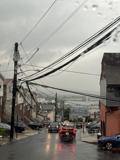 This image captures a street scene in North Bergen, USA, under a cloudy, overcast sky, suggesting inclement weather, possibly rain, as indicated by the water droplets on the lens and the wet street surface. The street is lined with residential buildings and is cluttered with a complex network of overhead power lines and utility poles, a prominent feature of the urban landscape. Several vehicles are present, including cars and trucks, some of which are in motion, with headlights and taillights illuminated, indicating traffic. The buildings appear to be multi-story, with some featuring brick or siding exteriors and visible staircases leading to entrances. The overall impression is of a somewhat densely populated urban area with aging infrastructure.
