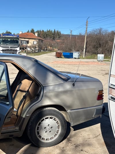 The image displays an outdoor scene in Baylovo, Bulgaria, under a clear blue sky with bright sunlight. In the foreground, the rear three-quarter view of a grey Mercedes-Benz sedan is parked on an unpaved, compacted dirt surface. The rear driver's side door is open, revealing tan fabric upholstery on the rear seat and a black seatbelt. The vehicle is equipped with 8-hole alloy wheels on the visible rear tire and features an antenna extending from the right rear fender. To the immediate right, a portion of a white caravan with horizontal brown and white striped panels is visible.
In the midground, a dirt lot contains a brown skip, a blue skip, and two silver wheeled waste bins. A large quantity of reddish, fragmented debris, possibly brick or tile, is scattered on the ground near these containers. A concrete utility pole with power lines extends vertically on the right side of this lot.
The background features several structures, including a two-story house with a red-tiled roof and white walls positioned behind a partially visible white Volvo truck on the left. The truck's cargo area is covered with a blue tarpaulin. Beyond the structures, a mix of coniferous and deciduous trees, primarily bare, covers a gentle incline, with hills visible in the distant background. No individuals are present in the scene.