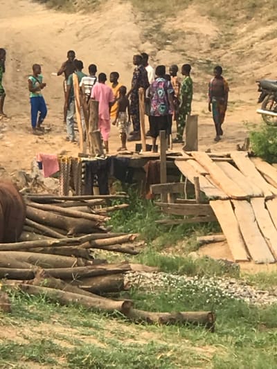 A row of people stand near a wooden structure and logs in a rural setting. Many of them wear colorful clothing. A dirt path runs beside them. To the side, a patch of grass grows near the wooden structure. A motorcycle is partially visible at the top of the frame.