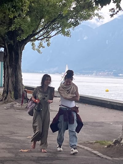 A daylight scene captured on a paved lakeside promenade in La Tour-de-Peilz, Switzerland. Two adult females walk side-by-side from left to right.
The individual on the left wears a loose, olive-green jumpsuit, carries a purple shoulder bag, and has a single pink sandal on her right foot while her left foot is bare. She holds a small, reddish-brown object with a white circular detail in her right hand and her left hand is open, palm up. She looks towards the second individual with a slight smile.
The individual on the right wears a white short-sleeved top, a beige scarf draped over her shoulders, light blue jeans, and a dark red garment tied around her waist. She has dark hair styled in a high bun with visible colored strands. Her arms are crossed, and her head is slightly bowed, looking towards the individual on the left. She wears white and black athletic shoes with visible Nike branding.
The environment includes a large tree with green foliage on the left, partially obscuring a light blue/teal structure and a brown bench. A concrete wall separates the promenade from a large body of water, likely Lake Geneva, where a white sailboat and a yellow buoy are visible in the distance. Hazy blue mountains rise in the background beneath a light blue sky.