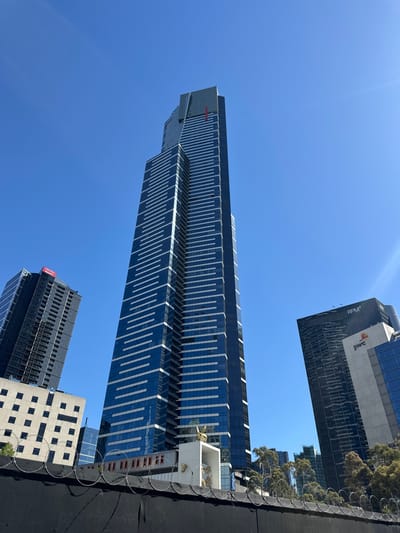 A tall skyscraper dominates the view against a bright blue sky. The building has a blue glass facade with repetitive white horizontal lines. Two smaller buildings are visible at the base, one dark and one lighter with a grid pattern. Another tall building is partially visible at the top with "PWC" printed on its side. Barbed wire runs horizontally along the bottom, obscuring the ground.
