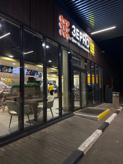 The image shows the exterior of a "Zerno" convenience store and cafe located in Maksimkovo, Russia, at night. The store features a modern design with large glass windows and a dark brown exterior facade. The "Zerno" logo, along with the slogan "для тех, кто в пути" (for those on the go), is prominently displayed in illuminated white lettering and orange circular shapes on the building's front.
Inside the store, visible through the windows, are dining tables with chairs, a service counter with a cashier, and displays of food and beverages. A woman in a yellow uniform is seen working behind the counter. Outside, there are brick-paved walkways and a paved road with black and white painted curbs. A trash can is situated to the right of the entrance. The overall scene suggests a well-lit and operational establishment catering to travelers or local residents during the evening hours.