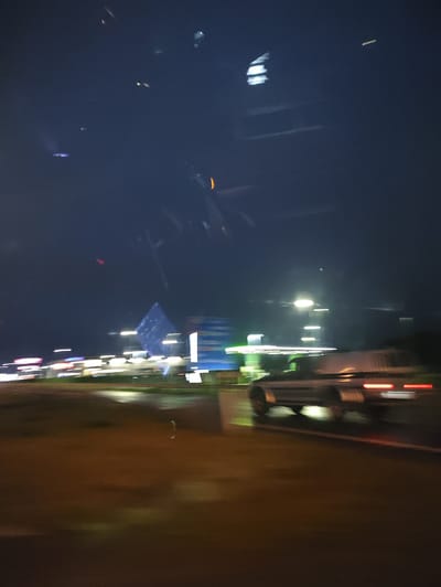 The image depicts a blurred night scene, likely taken from a moving vehicle, on a road in Laagri alevik, Estonia. The foreground is dominated by a wet, reflective surface, possibly asphalt with puddles, illuminated by the warm glow of streetlights and vehicle lights. In the mid-ground, a vehicle, possibly a pickup truck with a trailer, is visible, its taillights blurred red streaks, indicating movement. To the left, a row of buildings with illuminated signs and lights creates a blurry backdrop of commercial activity. Further to the left, a large, pyramid-like structure with blue lights is a prominent, albeit indistinct, landmark. The sky above is dark blue, with some faint streaks of light, possibly from other vehicles or reflections. The overall impression is one of a transient urban landscape at night, characterized by motion blur and the interplay of artificial lights against the dark sky.