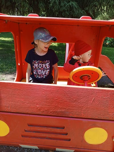 Two children are inside a red toy vehicle that resembles a truck or bus. The child on the left, wearing a black t-shirt with a floral design that says "MORE LOVE", a blue and grey cap, and glasses, is looking to their right with their mouth open. The child on the right, wearing a red hat and a red jacket, is holding onto a yellow and red steering wheel. The vehicle is outdoors, with green grass and trees visible in the background. The scene appears to be a playground or park in Verkhneyarkeyevo, Russia.