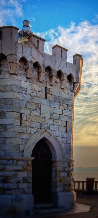 The image shows a close-up of the side of a lighthouse, made of stone blocks. The lighthouse has a distinctive architectural style with projecting elements and an arched opening. The sky above is blue with scattered clouds, suggesting it is daytime. The light indicates either morning or late afternoon. The weather appears clear. There are no people visible in the image, nor any explicit text. The scene suggests a coastal or elevated location, typical for a lighthouse.