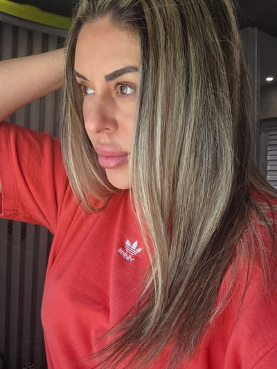 The image is a close-up portrait of a woman with blonde and brown highlighted hair, wearing a red Adidas t-shirt. She is looking to her left with her right arm raised behind her head. The background appears to be indoors with vertical blinds or paneling and some diffused lighting. There is no indication that the image was taken in Carpi, Italy.