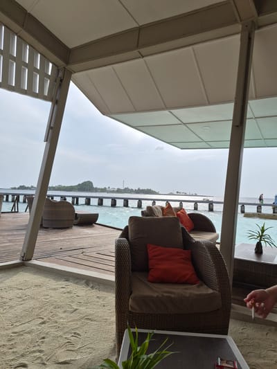 The image shows a partially covered outdoor seating area in Bodufolhudhoo, Maldives, looking out to the ocean. The seating includes wicker chairs with brown and orange cushions. A small table is next to the chairs, and a person's hand holding a lit cigarette is visible near the table. A long wooden pier extends into the turquoise water. The sky is overcast. A beach with sand and footprints is adjacent to the wooden deck where the seating is located.