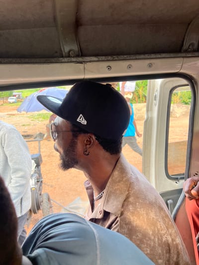 The image shows a side view of a man wearing a black baseball cap and sunglasses, looking out of a vehicle window. He has a beard and an earring. He is wearing a light brown collared shirt with a chain necklace. Another person in a light-colored jacket is partially visible on the left. Outside the window, a blue umbrella, some people, a car, and lush green vegetation are visible. The scene appears to be in a rural or semi-rural area, possibly Okwe, Nigeria, given the context. The lighting suggests it is daytime.