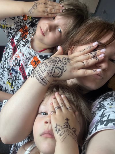In a dimly lit indoor environment, three individuals, presumably a mother and her two children, are captured in a close-up, selfie-style photograph. The subjects are adorned with henna-style temporary tattoos on their hands and arms, featuring intricate patterns and designs. The mother is positioned in the background, partially obscured by one of her children, with her hand raised to cover her eye, showcasing detailed henna art on her fingers and palm. Her lips are slightly puckered, and a ring with small stones is visible on her finger.
One child is in the foreground, with their face turned towards the camera and their hand covering their eye, also displaying henna tattoos on their fingers. The other child is positioned above and to the left, with their hand covering their eye and their face angled away from the camera, revealing henna markings on their hand and arm. All three individuals appear to be wearing patterned clothing. The background is nondescript, suggesting an indoor setting without specific identifying features related to the city of Аряж, Russia. The overall impression is a casual, playful moment shared among the family.
