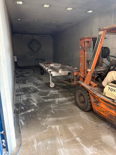 The image shows the interior of a dusty industrial workshop, likely in Mol, Belgium. An orange Toyota forklift is parked on the left side of the frame. The forklift has a person's arm resting on its console. In front of the forklift, a long, metal rack on wheels is positioned. The floor of the workshop is covered in a thick layer of white dust, with tire marks from the forklift clearly visible. The walls and ceiling are also dusty, and there are several square light fixtures on the ceiling. In the background, a diamond-shaped outline is visible on the wall. A blue hose runs along the floor, connected to a white electrical box on the left wall.