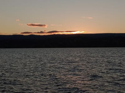 A wide, level shot captures the horizon over a body of water, presumably the San Roque Lake in Villa Carlos Paz, Argentina, given the setting sun and landscape. The sky is a gradient of orange and pink hues, obscured partially by a line of dark silhouetted mountains. Small, scattered clouds tinted pink by the setting sun hover above the mountains. The water surface is ruffled by a gentle breeze, reflecting the faint colors of the sky, though its overall tone is a deep, dark grey. No people or boats are visible in the frame.