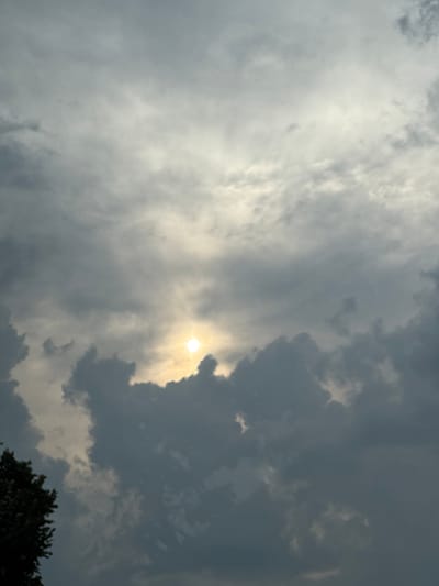 This is a landscape photograph taken outdoors, looking up at a dramatic sky filled with thick, grey clouds. The sun is partially visible through a break in the clouds, casting a bright, diffused light. The sunlight creates a luminous glow around the sun, illuminating the edges of the surrounding clouds and creating a sense of depth and texture. The clouds vary in darkness, with some appearing almost black and others a lighter grey, suggesting a dynamic and possibly stormy weather system. In the lower right corner of the image, the dark silhouette of the top of a leafy tree is visible, providing a grounded element to the otherwise ethereal sky. The overall mood is atmospheric and somewhat moody, with the interplay of light and shadow in the clouds being the main focus. The time of day is not explicitly stated, but the quality of light suggests it could be late afternoon or early evening, possibly before or after a storm. There is no visible text in the image.