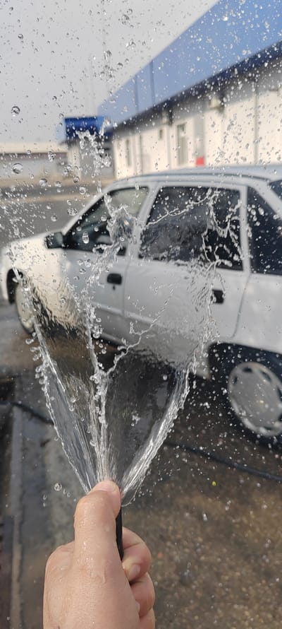 A blurry white car sits in the background, partially obscured by a hand holding a black hose that sprays water. The water droplets are scattered across the foreground and around the car, creating a dynamic, splashy effect. A building with blue and white trim is visible in the distance, also slightly out of focus due to the water spray. The scene suggests a car wash or an attempt to clean the car with a hose.