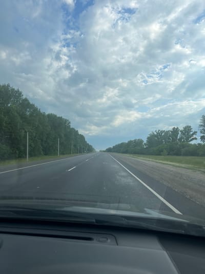 The media file depicts a view from inside a vehicle driving on a paved road. The road has a white line marking the lane. On the left side of the road, there is a dense line of green trees. The sky is filled with white and gray clouds, indicating overcast weather. On the right side of the frame, the interior of the vehicle is visible, showing dark gray panels and a white line that appears to be part of the car's interior design. The scene suggests a journey through a rural or suburban area. The time of day appears to be daytime, given the brightness of the sky. There is no visible text in the image.