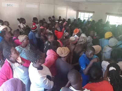 This media file captures a crowded indoor gathering of predominantly women in what appears to be a community hall or meeting room in Butali Chegulo ward, Kenya. A large group of people, mainly adults of African descent, are densely packed within the space, either seated or standing. Many individuals are visible, often with their heads and upper bodies.

The subjects are dressed in various forms of casual attire, including brightly colored clothes and head coverings such as scarves and wraps worn by many of the women. Their attention appears to be directed towards a common focal point, presumably a speaker or presenter, indicating they are participants in a meeting, workshop, or community event. Their expressions are generally attentive and engaged.

Natural daylight streams into the room through windows visible in the background, suggesting the event is taking place during the daytime. The room features plain, light-colored walls, indicative of a functional public or community space.

Visible text on the walls includes "SEC." clearly displayed on the left, likely an abbreviation for 'Secretary' or 'Section'. Another less distinct text that appears to read "M I Z" is visible higher up on the right wall. The overall scene conveys a strong sense of community engagement and shared focus during an organized gathering.