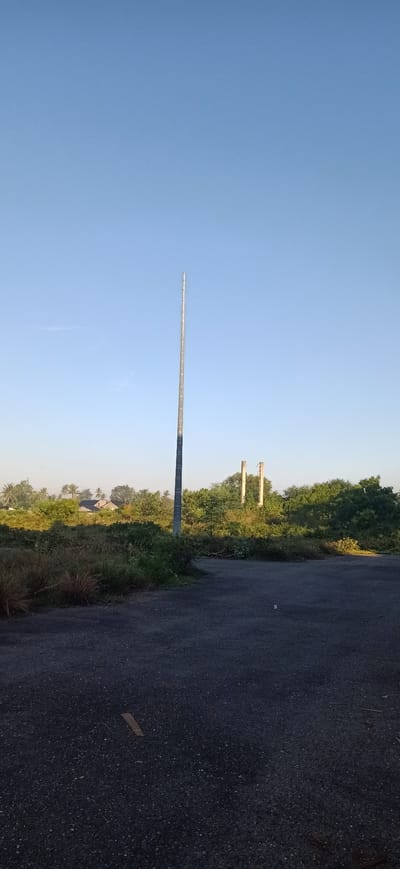 In Uteungeulinggang, Indonesia, a tall, slender, silver-colored pole stands vertically in the center of the image, reaching towards a clear, pale blue sky. The pole appears to be made of metal and has a textured surface with horizontal markings. The foreground is dominated by a dark asphalt surface, with sparse patches of dry grass and debris scattered across it. To the left of the pole, a dense area of green foliage and tall grasses creates a natural border. In the background, beyond the pole and foliage, a line of lush green trees and palm trees stretches across the horizon. Two unfinished concrete pillars, weathered and rough, are visible amidst the greenery to the right of the main pole. In the distance, several houses with dark roofs peek through the trees. The overall scene suggests an outdoor, undeveloped area, possibly on the outskirts of a residential zone, bathed in the soft light of either early morning or late afternoon. There are no people or animals visible in the frame.