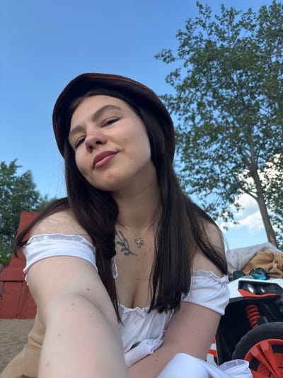 A woman wearing a brown hat and a white top is pictured outdoors, possibly in Пальники, Russia. The background includes a blue sky, green trees, a red structure, and what appears to be a toy car. The woman is looking at the camera with her tongue slightly out. She has a tattoo on her chest.