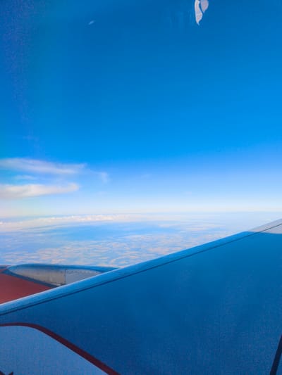The image depicts an aerial view of the landscape, likely from an airplane window. The sky is a vibrant, clear blue, with a few wispy clouds near the horizon. Below, the Earth is visible, a patchwork of fields and possibly some sparsely populated areas, suggesting a rural or semi-rural setting. The colors of the landscape are muted, with shades of brown, green, and beige indicating agricultural land. The edge of an airplane wing, painted in shades of blue and red, occupies the foreground, indicating the vantage point of the photograph. The wing appears to be covered in a fine texture, possibly condensation or dirt. There are no visible people or specific structures that would pinpoint a location within Sallgast, Germany without further context. The scene is tranquil and vast, emphasizing the expansive nature of the sky and the distant, observable land.