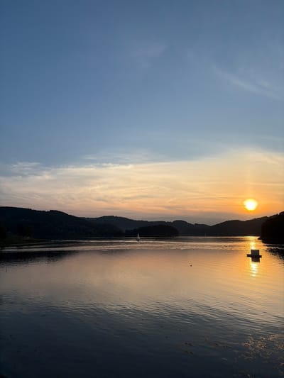 This is a vertical, eye-level, medium shot of a lake at sunset, with a mountain range in the background. The sky is a gradient of blue to orange, with wispy clouds. The sun is setting over the mountains, casting a golden reflection on the water. A sailboat is visible in the distance on the left side of the lake. On the right side of the lake, a small, white structure floats on the water, reflecting the sunset. The water is calm, with gentle ripples. The foreground of the image is dark, with the water appearing almost black. The mountains in the background are silhouetted against the bright sky. The scene appears to be set in Gródek nad Dunajcem, Poland, given the characteristic landscape of the area. There are no people or animals visible in the image.