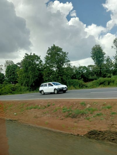 A white car travels along a paved road. Lush green trees line the roadside, contrasting with a strip of brown earth and sparse vegetation. Above, a partly cloudy sky fills the frame, the clouds exhibiting shades of gray and white with hints of blue sky visible. The foreground is out of focus.