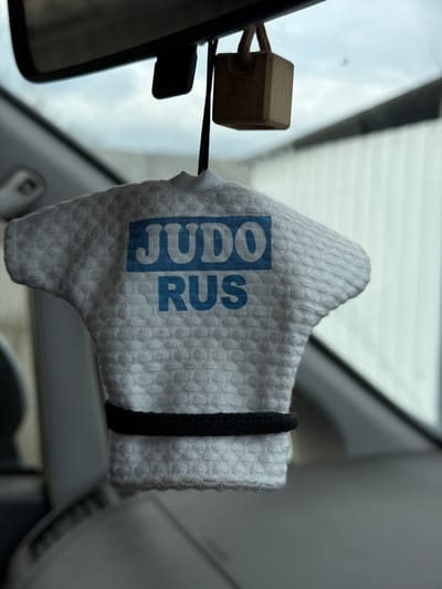 A car interior is seen, with a rear-view mirror visible on the left. Hanging from the mirror is a light-colored, textured object shaped like a judo gi, emblazoned with the text "JUDO RUS" in blue. A black elastic band encircles the upper portion of the "gi." A small, unfinished wooden cube hangs near the mirror, suspended by a thin black cord. The background shows parts of the car's windshield, roof, and window frame. The image does not contain information to confirm the location in the city of Расцвет, Russia.