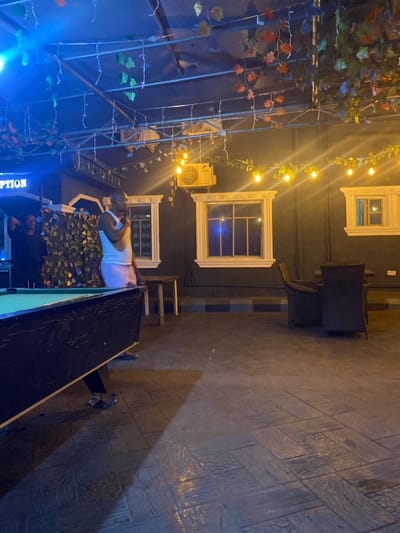 The scene depicts an outdoor or semi-enclosed leisure area at night. The specific location within Igbessa, Nigeria, is not discernible from the image. In the left foreground, a dark-framed pool table with a green felt surface is present. Two males are visible; one light-skinned male, wearing a white tank top and light-colored shorts, stands mid-frame near the pool table, facing right with his right hand near his chest. Behind the pool table, a darker-skinned male in a black t-shirt stands near a structure adorned with artificial foliage and a blue neon sign partially displaying "PTION." The background features a dark wall containing three white-framed, grid-patterned window-like structures. A white air conditioning unit is mounted above the central window structure. Overhead, a metal truss structure supports multiple strands of warm yellow incandescent string lights and garlands of artificial leaves. The floor is dark concrete, patterned to resemble wooden planks. To the right, a dark wicker table with two matching chairs is positioned. A small, dark wooden table stands between the pool table and the wicker seating. A blue and grey striped curb runs along the base of the dark wall.