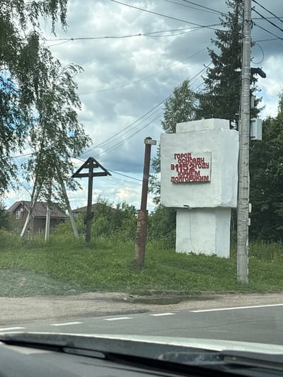 This media file shows a stone monument with red Cyrillic text on it, located in Pereslavl-Zalessky, Russia. The monument appears to be a historical marker, possibly indicating the founding date of the town or a significant event. The text, though partially obscured and viewed at an angle, reads: "ГОРОД ОСНОВАН В 1152 ГОДУ ПРИ ИГОРЕМ ДОЛГОРУКИМ" which translates to "The city was founded in 1152 by Igor Dolgorukiy".
The monument is situated in a grassy area next to a road, suggesting it is a roadside attraction or landmark. In the background, trees and power lines are visible, with a cloudy sky overhead, indicating overcast weather. A wooden cross, also a significant religious or historical symbol, is partially visible to the left of the monument. The overall scene suggests an outdoor setting, possibly in a rural or suburban area, and the monument itself is a prominent object. The image appears to be taken from a vehicle, as evidenced by the car interior visible on the right side of the frame. There is no indication of specific time of day or emotions conveyed.
