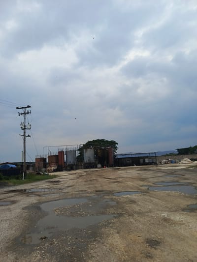 The image shows an industrial area in El Bajío, Venezuela, under a cloudy sky. There are several storage tanks and an electrical pole. The ground is dirt with puddles of water. In the background, a structure with piles of materials can be seen. There are no people visible in the frame.