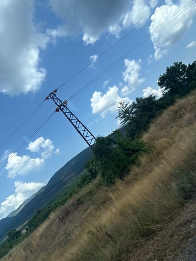 The image, captured in the vicinity of Trapoklovo, Bulgaria, presents a high-angle, upward-looking perspective of a power line tower and its associated cables against a bright blue sky dotted with fluffy white clouds. The tower, a skeletal metal structure, is positioned on the right side of the frame, angled diagonally upwards and to the left. It supports multiple dark power lines that stretch across the upper portion of the image.
The foreground and middle ground are dominated by a steep, dry, grassy slope rendered in shades of golden brown and ochre, indicating a likely summer or late autumn season. Scattered amidst the dry grass are patches of green vegetation and trees, which become denser and darker further up the slope, transitioning into a lush green hillside in the background. The distant background features the undulating contours of a mountain range, its slopes covered in a mix of green vegetation and darker shadows. The overall impression is one of a rural, possibly mountainous landscape under a clear, sunny sky.