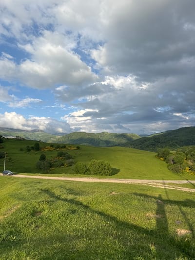 The image depicts a vast, green, rolling landscape under a dramatic sky filled with both white and dark grey clouds. In the foreground, a grassy slope transitions into a dirt road that curves through the middle ground. A dark-colored car is parked on the left side of the road. The mid-ground features more lush green hills dotted with trees and patches of yellow flowering bushes. In the background, a series of layered, tree-covered mountains recede into the distance, with some of the peaks partially obscured by clouds. The lighting suggests it is late afternoon or early evening, with shadows cast across the foreground grass. The scene is one of natural beauty and apparent tranquility, likely in a rural or mountainous region such as the area around Nizhnyaya Mara, Russia.
