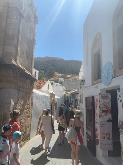 A group of people are walking down a narrow, sun-drenched cobblestone street in Lindos, Greece. The street is flanked by whitewashed buildings with traditional architecture. On the left, a large stone building with arched openings dominates the foreground. On the right, a building with a bright blue circular sign that reads "Rovo" features an advertisement for "Sundaes Creation" and "Bubble Tea." Several individuals, including children, are visible, dressed in casual summer attire. The sun casts long shadows on the ground. In the background, the Acropolis of Lindos and the surrounding ancient fortifications are visible on a rocky hilltop under a clear blue sky.