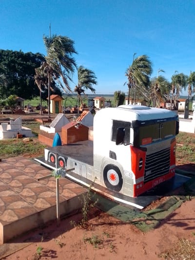 The image shows a cemetery in São Gabriel Do Oeste, Brazil. A prominent feature is a tomb shaped like a truck, painted white and red with the name "SCANIA" visible on the grille. The truck tomb is situated on a flat platform. In the foreground, a simple wooden cross with some artificial flowers is planted in the reddish-brown soil. Several other graves and small structures, typical of a cemetery, are visible in the background, along with palm trees swaying in the wind and a clear blue sky. The overall impression is that of a sunny day in a rural cemetery.