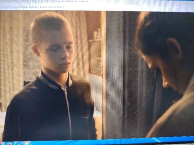 The image is a screenshot of what appears to be a scene from a film or television show, captured on a computer screen.
In the foreground and occupying the left side of the frame is a young person, likely a teenager, with a very short, buzz-cut hairstyle. Their head is slightly tilted down, and their gaze is directed downwards, suggesting a thoughtful, solemn, or perhaps withdrawn emotional state. They have fair skin, and there are what appear to be smudges or marks on their face, particularly noticeable on the nose and cheek. They are wearing a dark, possibly navy or black, bomber-style jacket with a distinctive collar featuring thin white horizontal stripes. The jacket is zipped up.
On the right side of the frame, partially obscured and facing away from the viewer, is a second person. Only the back of their head and upper shoulder are visible. Their hair appears dark and is either short or pulled back. They are also looking downwards, mirroring the posture of the person on the left, but their face is not visible.
The setting appears to be indoors. Behind the first person on the left, there's a textured background that could be patterned wallpaper or a curtain in a muted, light-colored tone. Between the two individuals, a vertical structure, possibly a doorframe or the edge of a wall, divides the space. The lighting in the scene is somewhat dim and naturalistic, casting subtle shadows and highlighting the subjects without being overly bright. The overall mood conveyed by the image is one of quiet contemplation or a moment of serious interaction.
Text from the computer screen interface is visible at the top and bottom of the image. At the top, there is a string of characters that resembles a file path or URL for media playback: "seodo playorida3c/33986-dbe5-4214-87-17-460806.50 10.vo-poltsana son-i-". Below this, there is Russian text that translates to "neither Windows 10 nor later versions. You have Windows 7 installed," indicating the operating system of the computer on which the screenshot was taken. At the very bottom of the image, a blue bar, characteristic of a Windows taskbar or application window border, is visible.